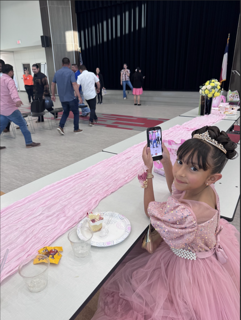 A girl in a pink tutu dress takes a selfie at a table. Behind her, people walk and sit.