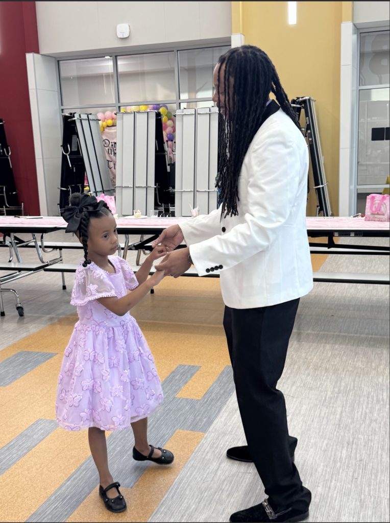 Man in white suit holding hands with a young girl in pink dress and black shoes, in a room.