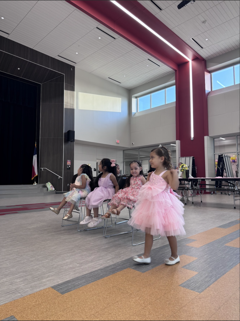 A few girls sit in chairs in a room with a stage in the background. One girl stands in front of them.