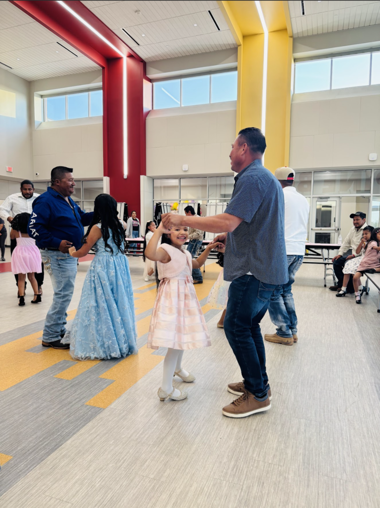 Two children dance with adults in a large room. The children wear dresses, and the adults wear casual clothes.