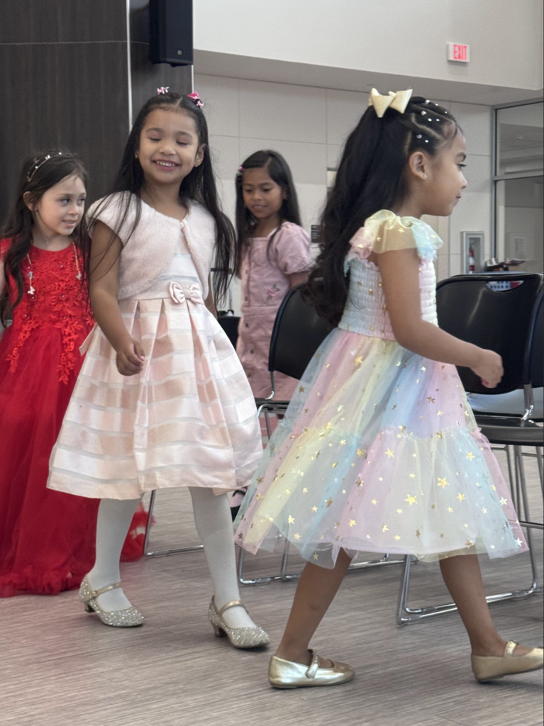 Four girls in various dresses walk in a hallway, one with a red dress and two with pink dresses.
