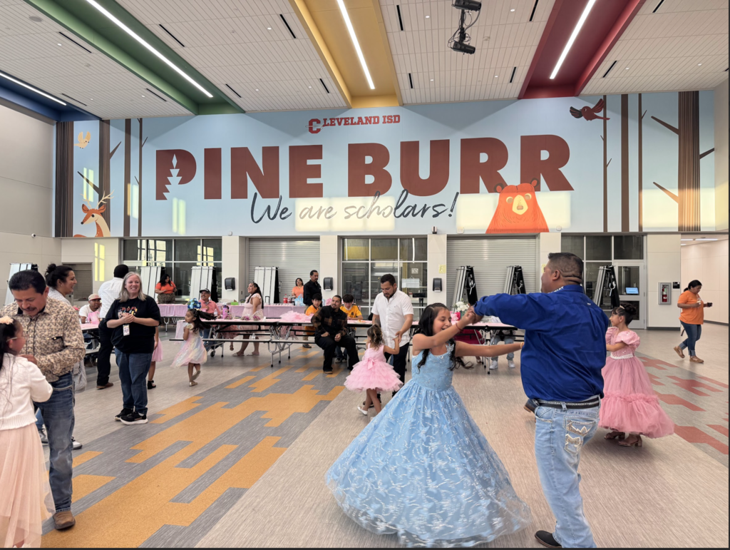 A group of people, including children and adults, dance in a hallway with the banner "PINE BURR" in the background.