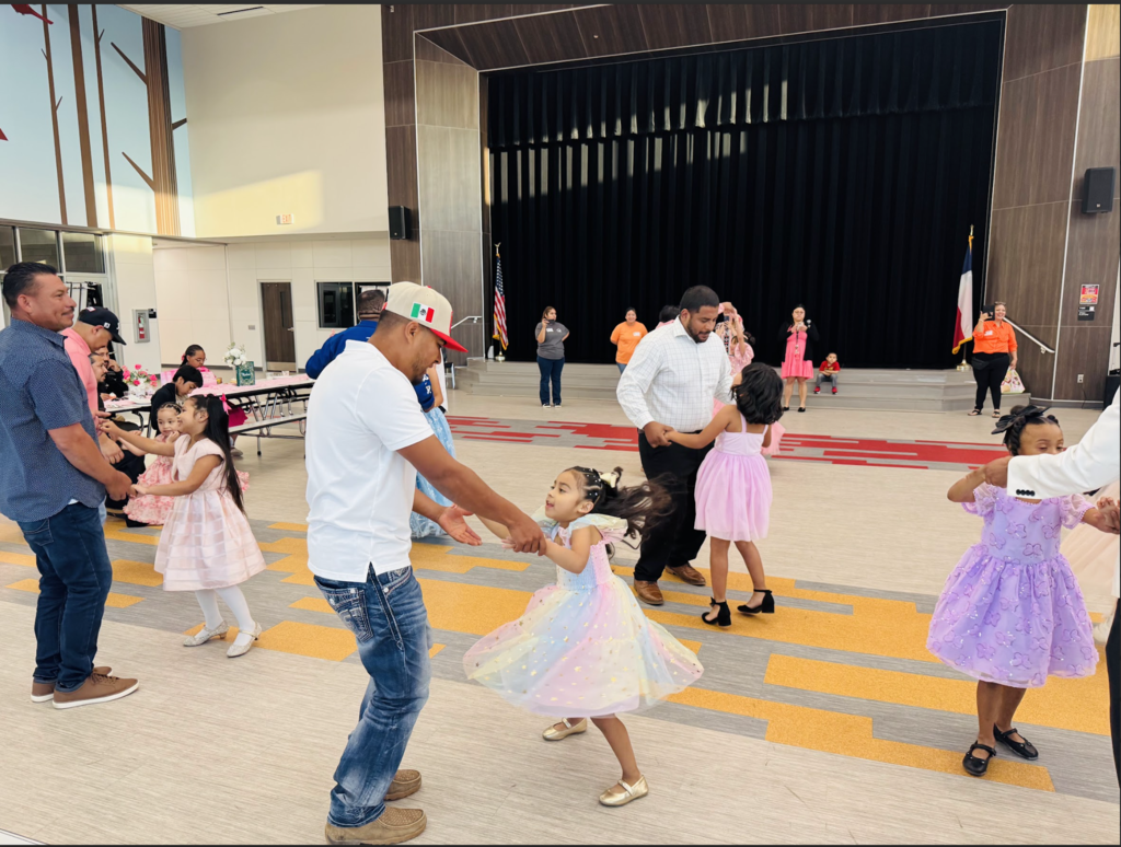 Group of people dancing in an auditorium, including a man with a baseball cap and a girl in a pink dress.