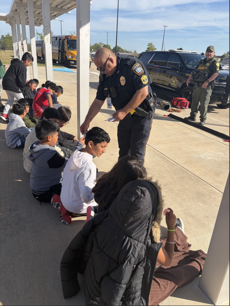 Police officer checks on seated children while another officer stands behind them, with a vehicle and trees in the background.