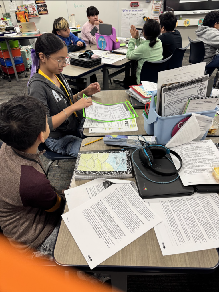 Children seated at tables in a classroom. One child looks at another child. Papers, books, and a map are on the table.
