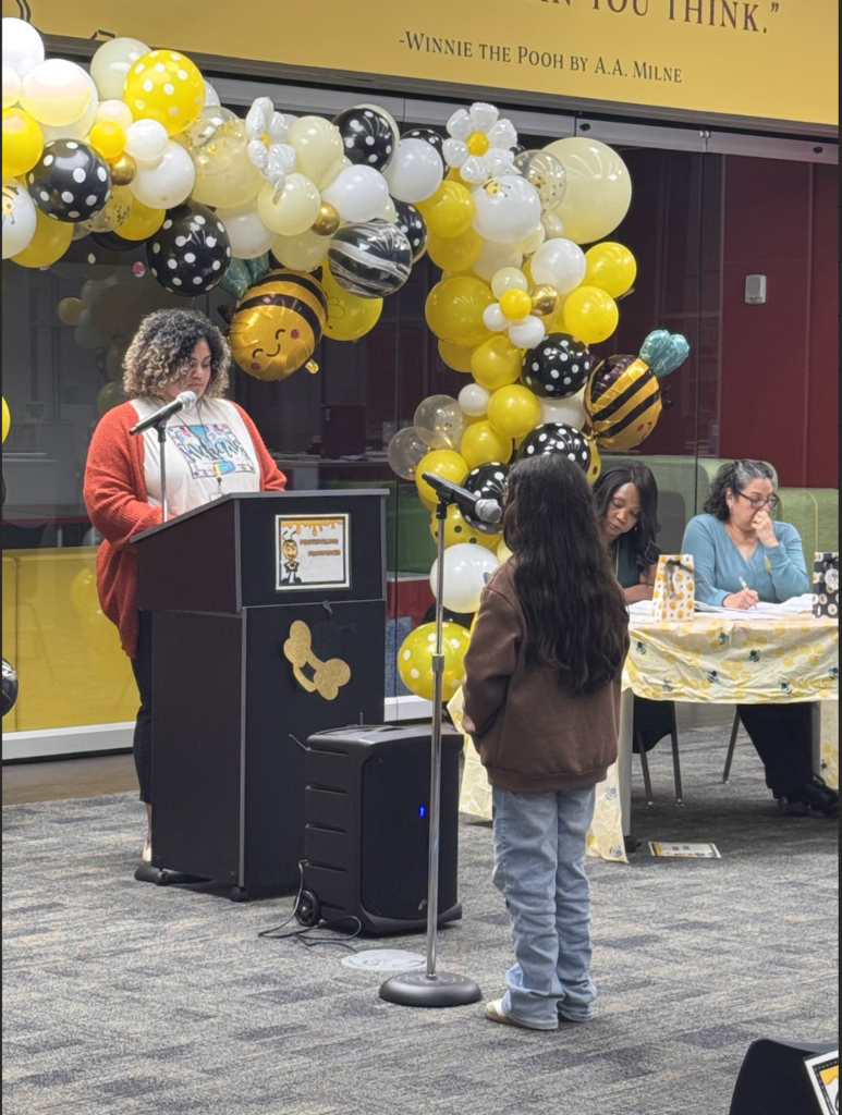 Person at podium with microphone, a child stands close, and two people sit at a table. Behind them, balloons form a colorful arch.