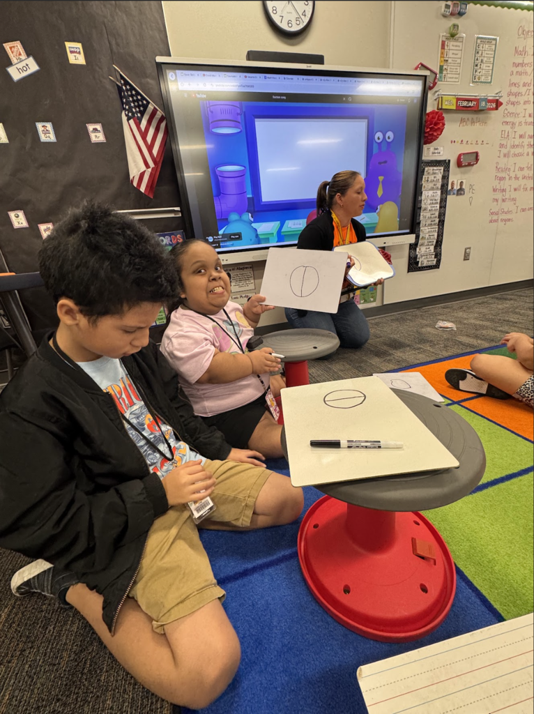 Children seated at a table, engaged in an activity with a large monitor and teacher present in the background.
