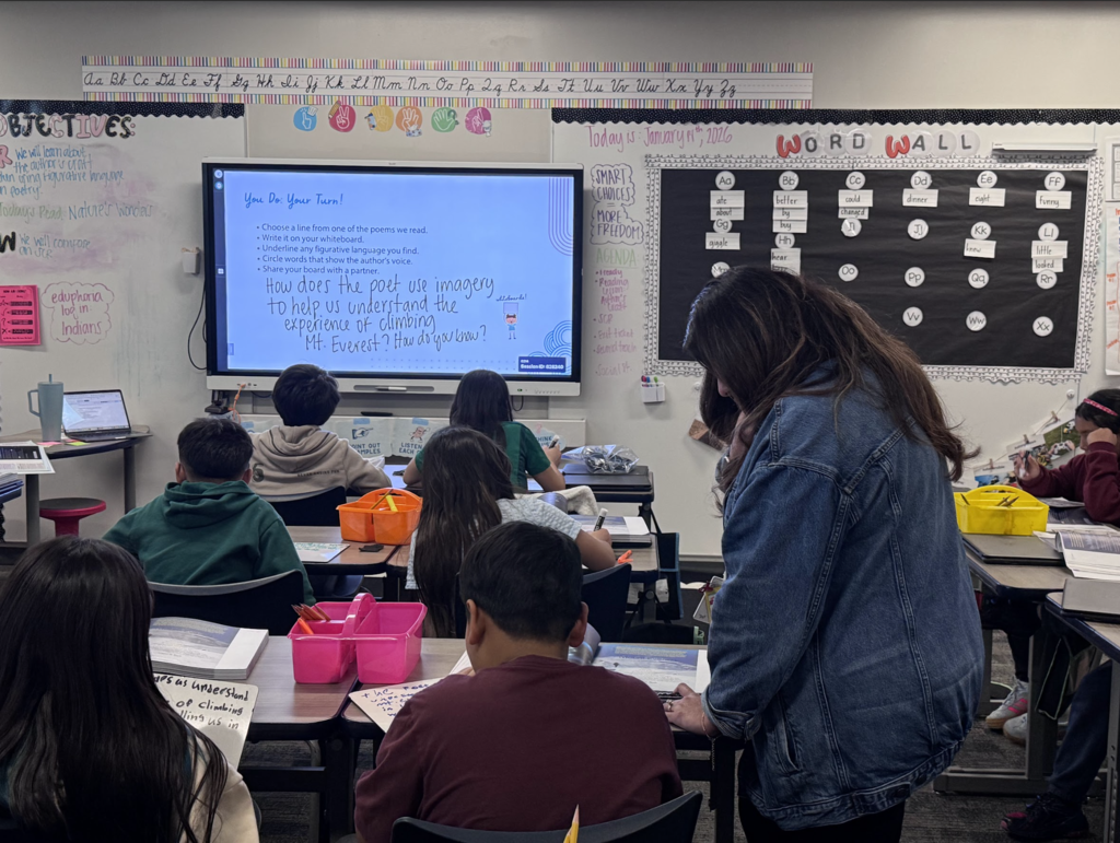 Classroom with students seated at desks. An instructor stands by the front wall with a whiteboard and screen.