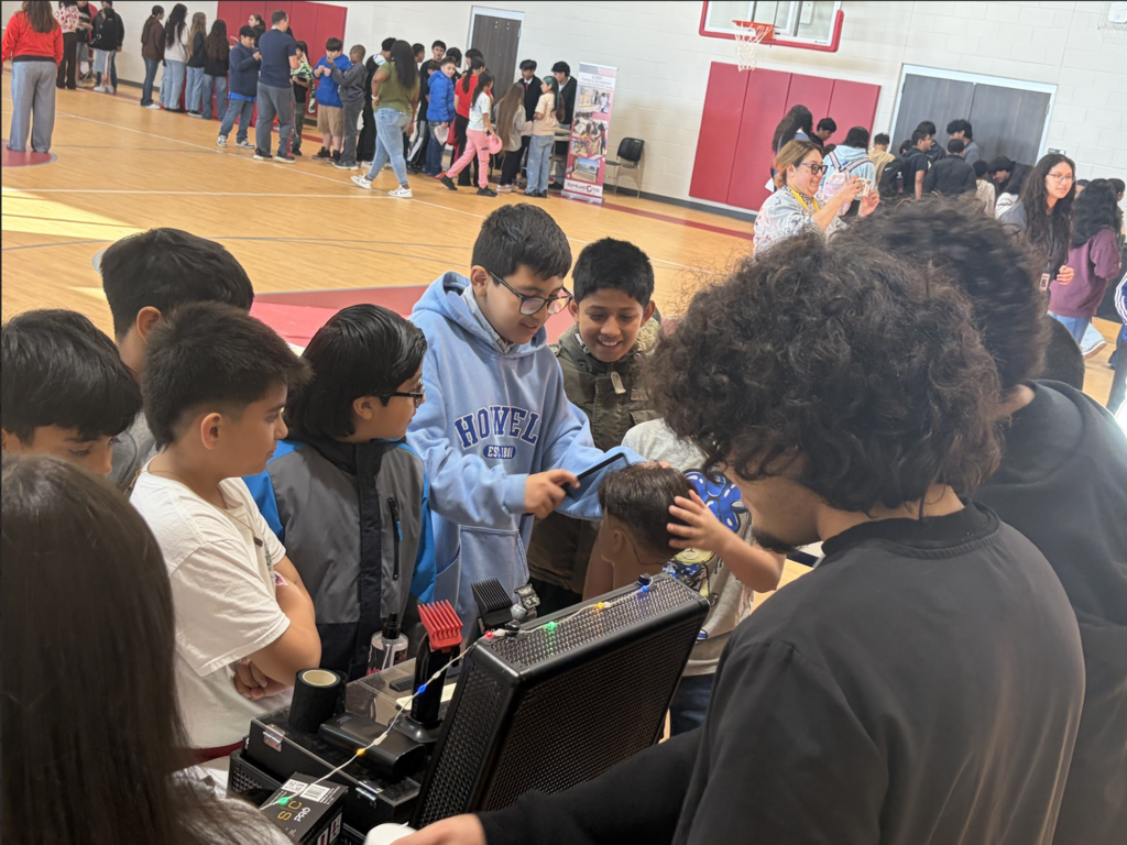 A group of students gather around a computer monitor, engaged in a project. An adult helps a child behind them.