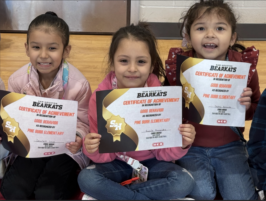 Three girls seated on a floor, holding certificates, with one wearing pink, one in red, and the other in blue.