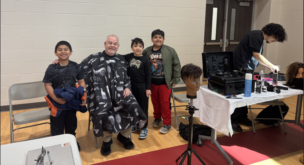 Five individuals in a gym. One person has a cape around their neck. Another person works on a mannequin head.