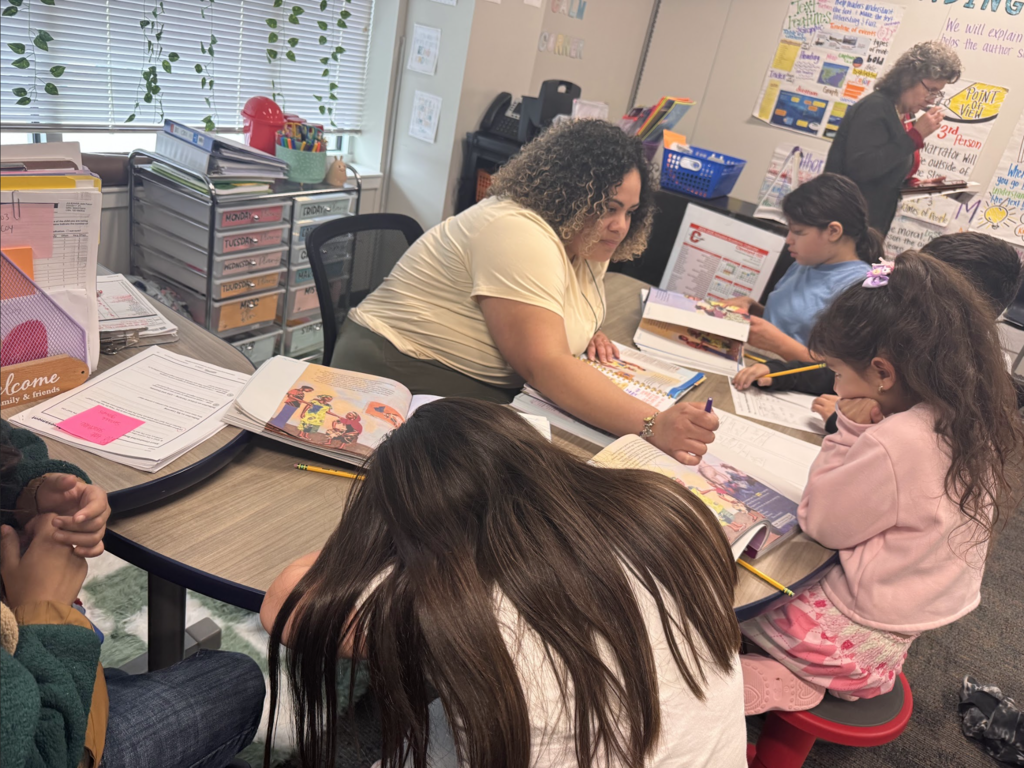 A woman assists children at a table. They are seated around the table, each working on books and papers.