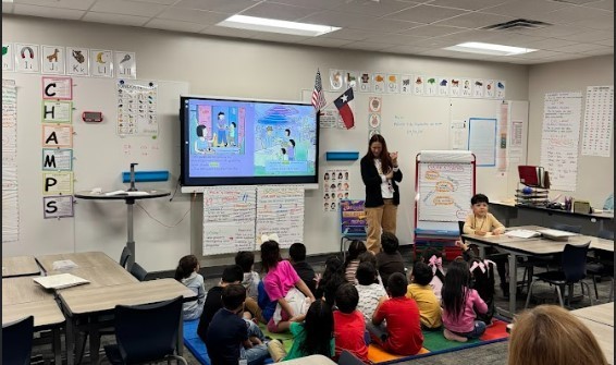 A teacher shows a presentation to young students sitting on the floor in a classroom.