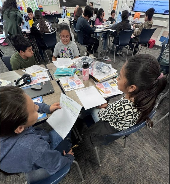 Group of students in a classroom; some seated at desks, others standing. Papers and books on desks.