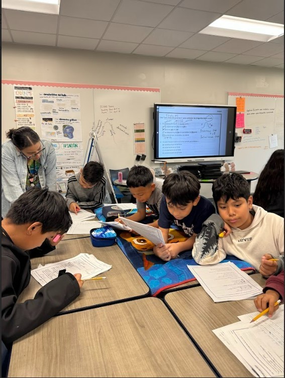 Students are seated around a table, engaged in reading and writing activities. A teacher stands behind them.
