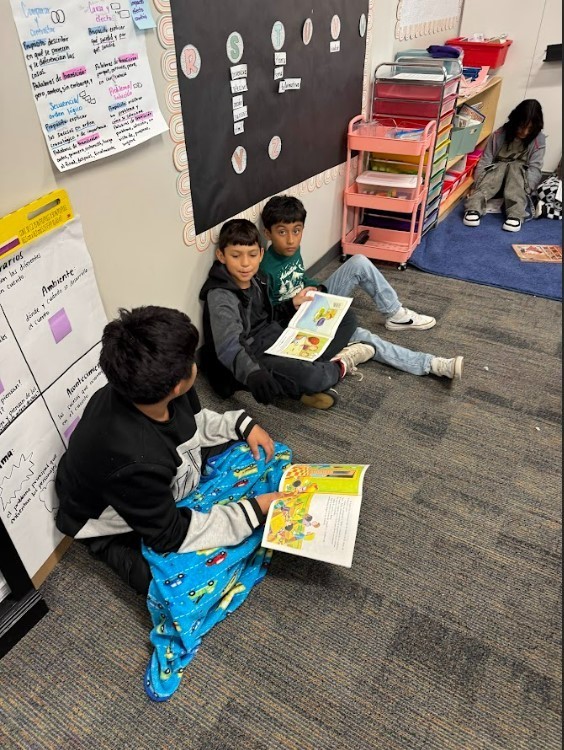 Three children sit on the floor reading books, with a bulletin board, shelf, and another child behind them.