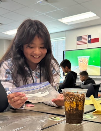 A person with long hair examines a plastic bag at a desk, next to a drink.