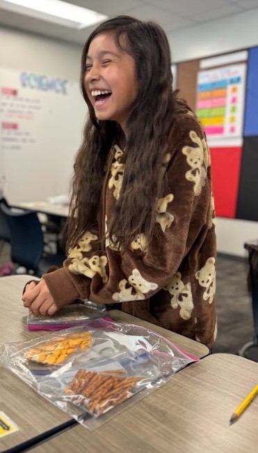 Girl standing in classroom with brown coat, smiling, surrounded by snacks on a table in front of her.