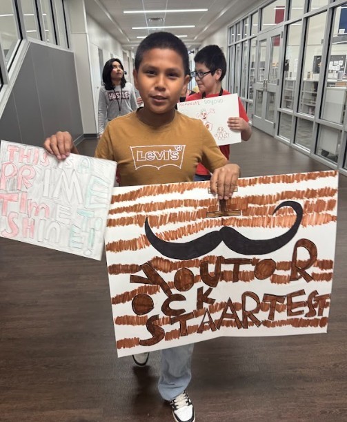 A young boy holds up signs promoting an event. The signs feature mustaches and text. Another child holds a sign behind him.