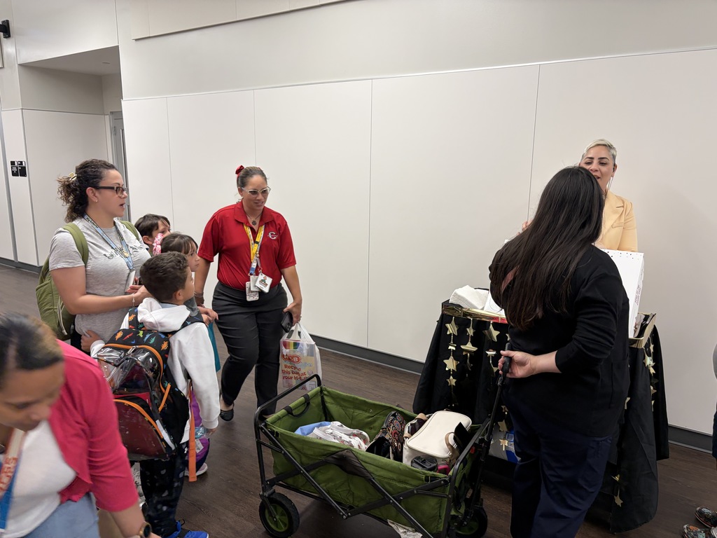 People stand in a room with white walls, some carrying backpacks. A woman pushes a cart with items.