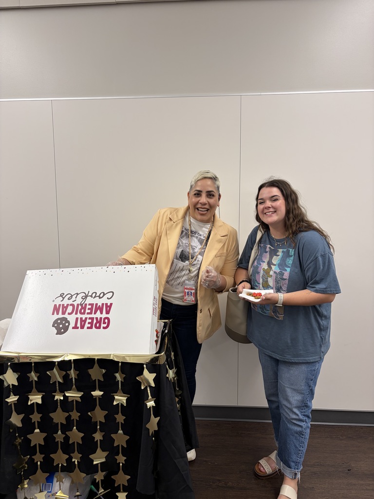 Two women stand by a table with a box labeled "Certified Real Deal" and a decorative tablecloth. One holds a pizza.