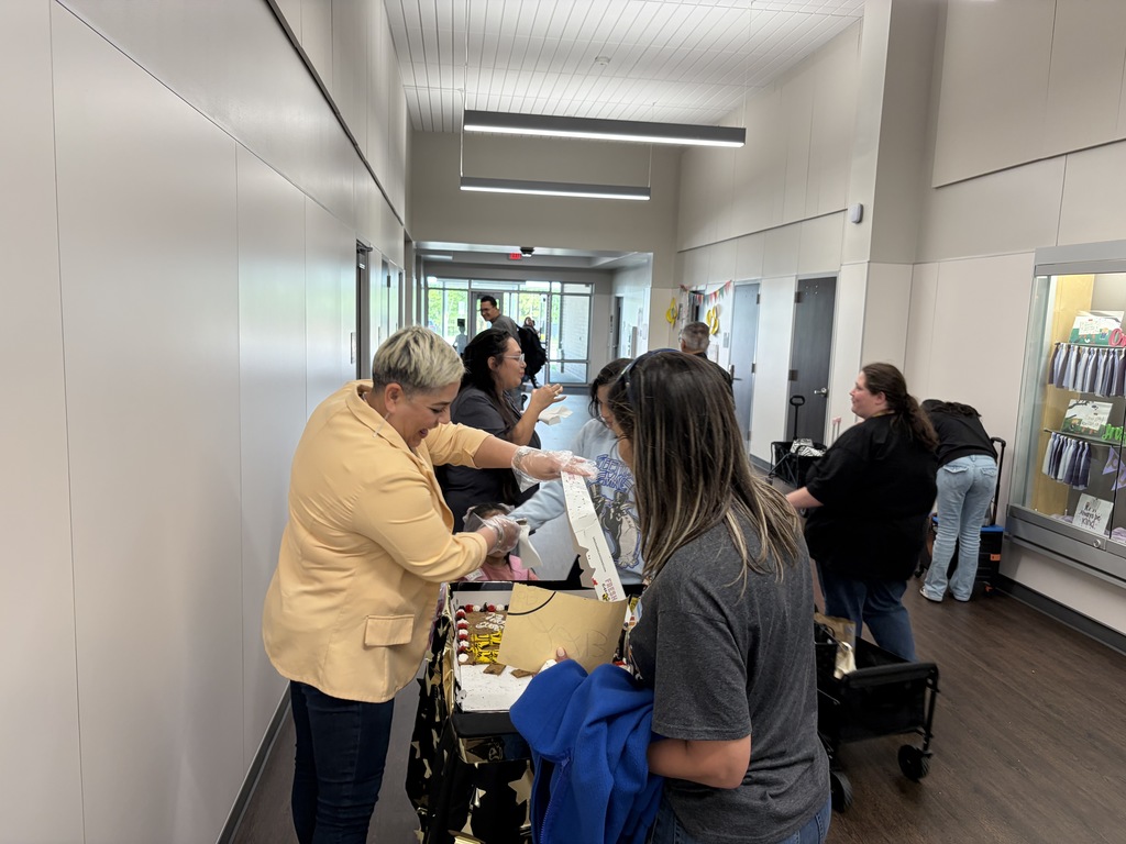 Several people are standing in a hallway, with one woman in a yellow blazer reaching into a box.