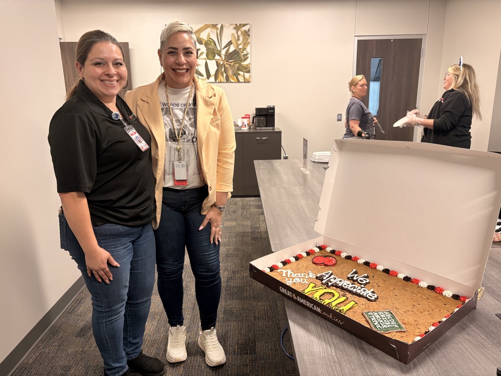 Two women stand next to a cake in a box. One wears black. The cake has words. Others are in the background.