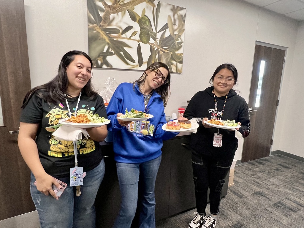 Three women, each holding a plate of food, stand next to a counter. Behind them is a wall with a painting.
