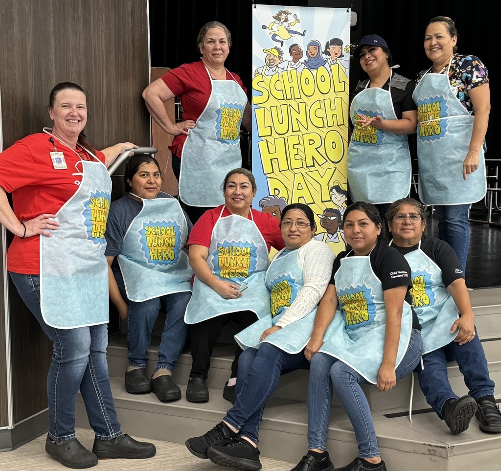 A group of women wearing aprons pose on a stage with a banner reading "SCHOOL LUNCH HERO DAY".