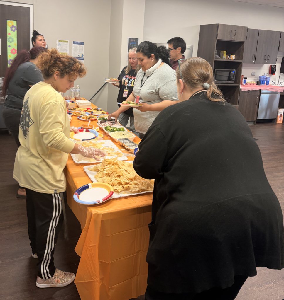 People stand around a long table with food on it in a room with cabinets and a microwave.