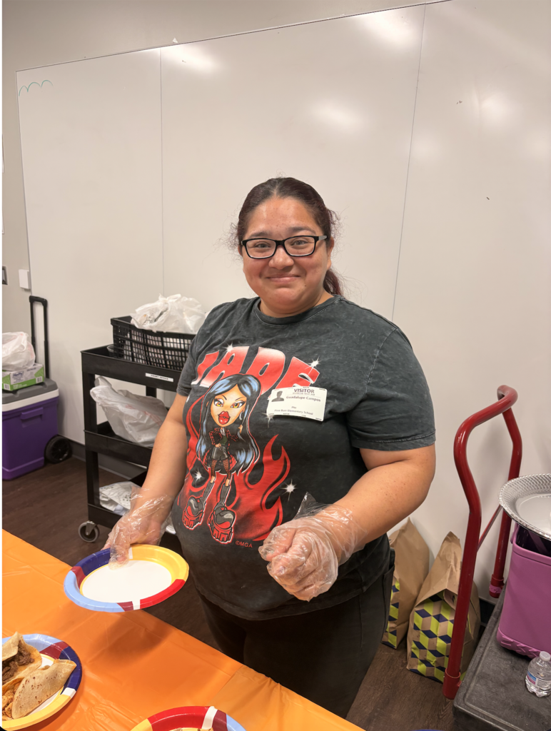 Woman in a gray t-shirt holds a colorful plate with food, smiling. Background includes a white wall and a cart with items.