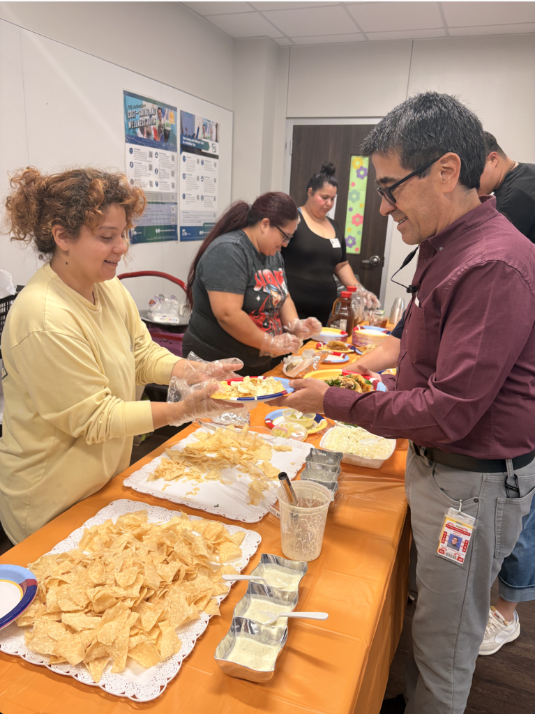 People stand at a table serving food; a woman in yellow holds food, and a man in maroon reaches for the food.