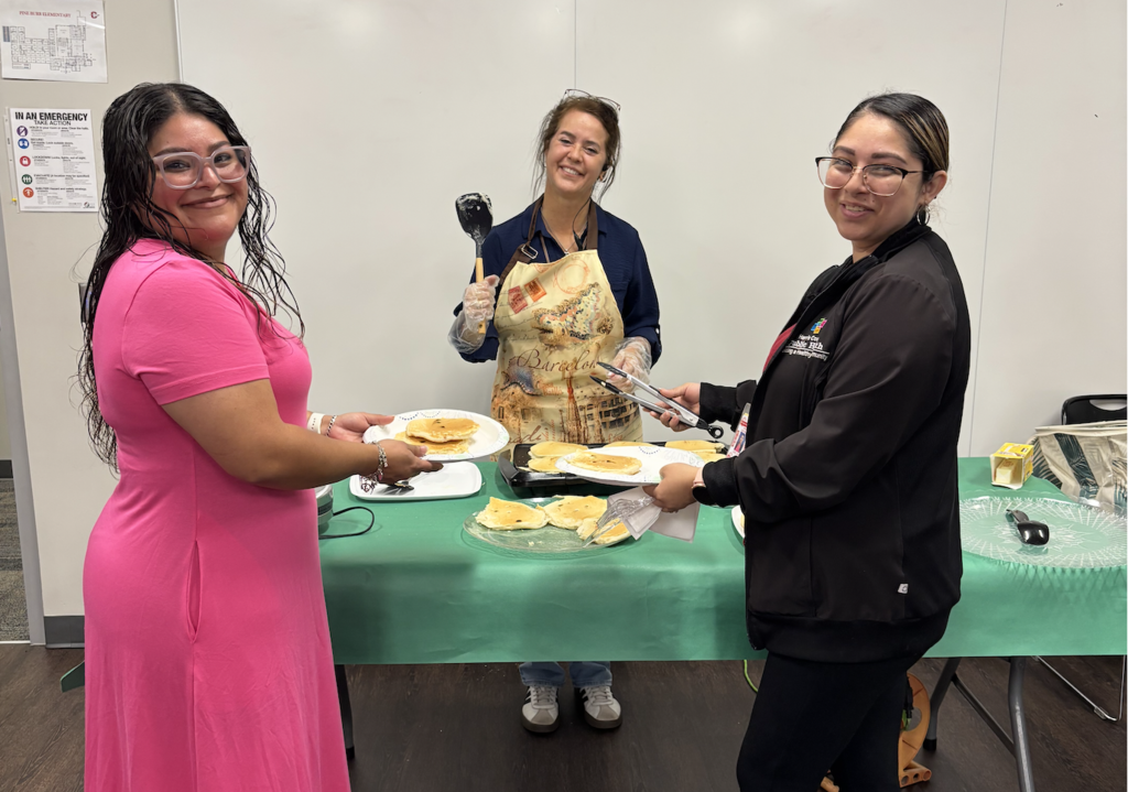 Three women in a room. One in a pink dress holds a plate, another in an apron holds utensils, and one in black holds a plate.