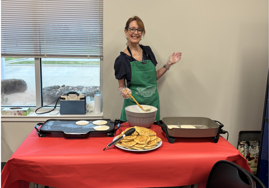 A person in a green apron stands behind a table, mixing ingredients in a bowl, with cooked pancakes on the table.