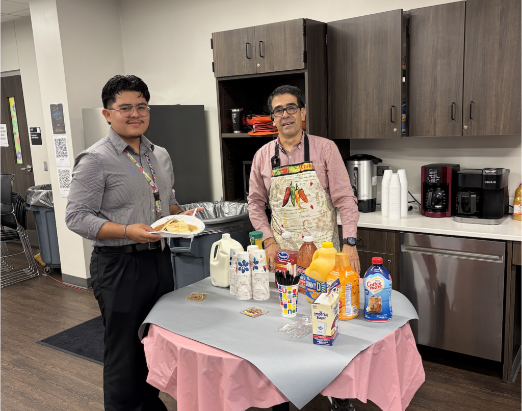 Two people stand in a kitchen, one wearing an apron, holding a plate of food. A table has various beverages and food items on it.