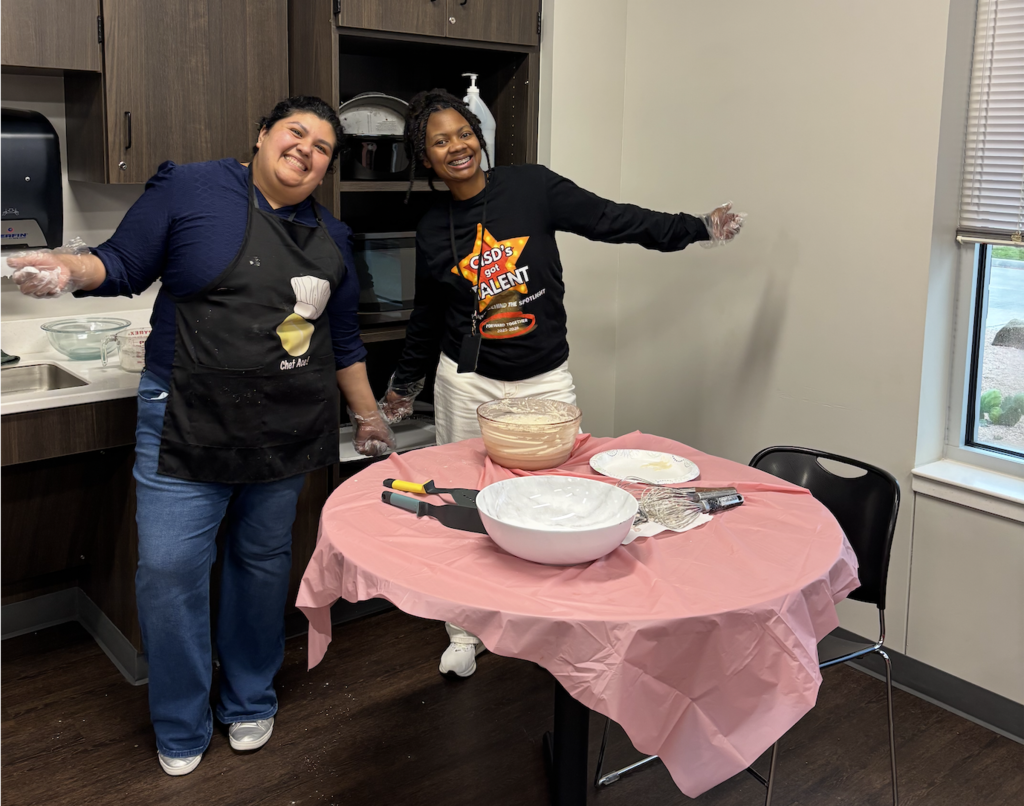 Two people in aprons, standing in a kitchen, one with hands raised, next to a table with a pink cloth.