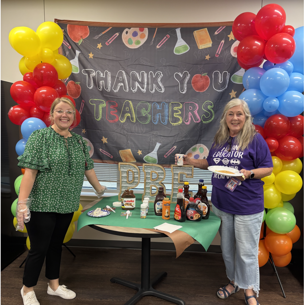 Two women standing by a table, one holding a plate of food, near a banner reading "Thank You Teachers".
