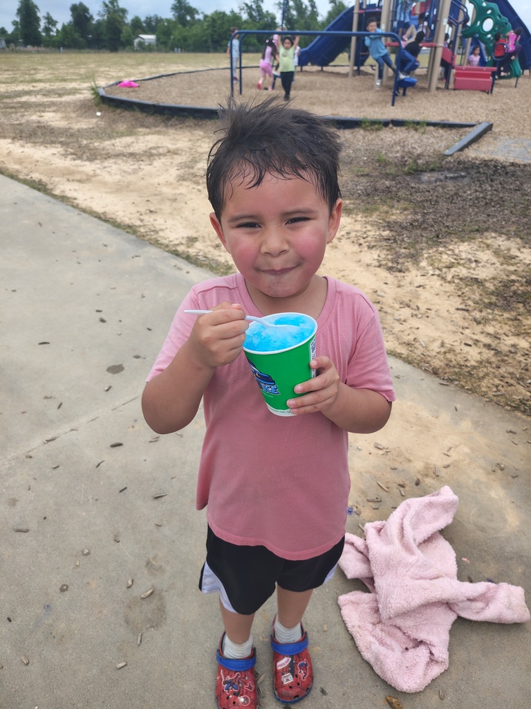 Child in pink shirt and shorts holds a blue cup of frozen beverage on a sidewalk with playground behind.