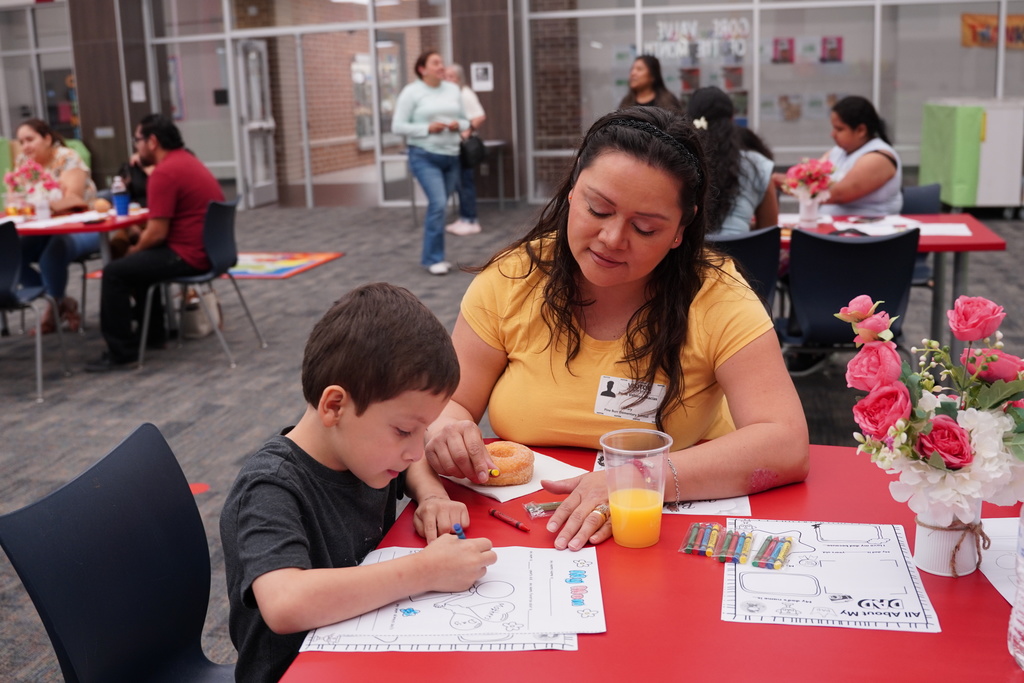 A mother and child are seated at a table with food, a flower vase, and coloring pages.