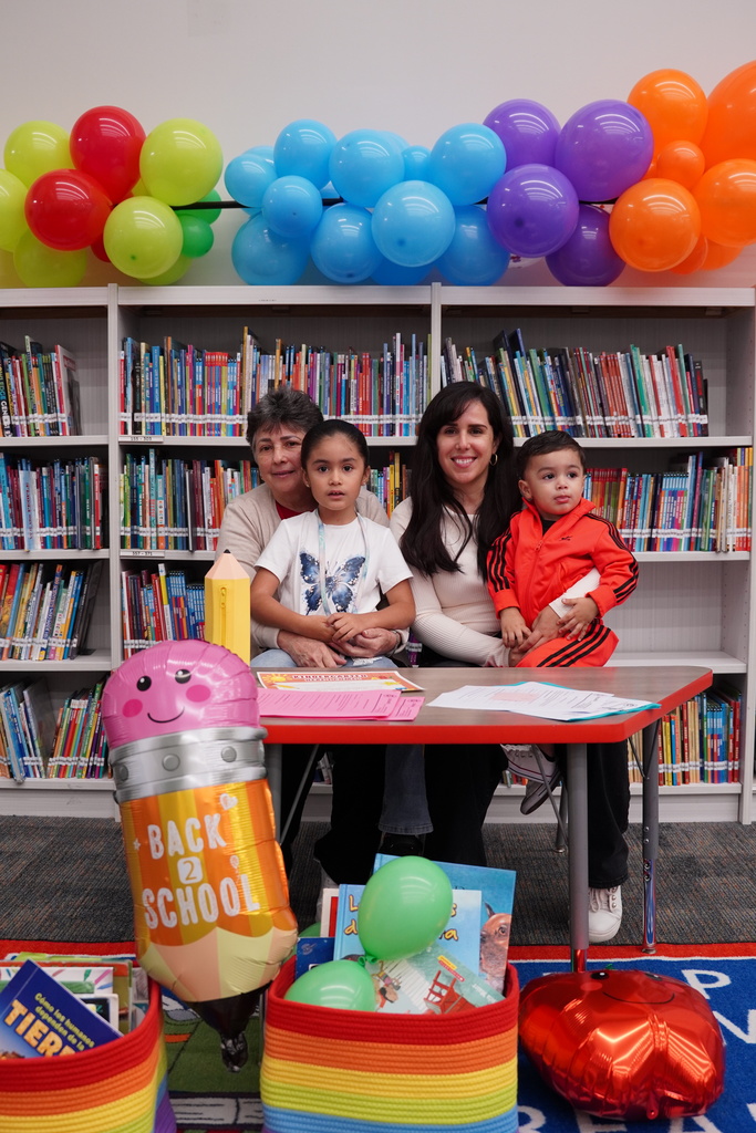 A family of four, seated at a table with books and balloons in the background, is smiling for a picture.