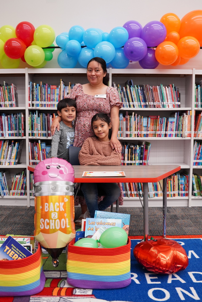 A woman and two children sit at a table with a large balloon. They are in a library with shelves of books and colorful balloons.