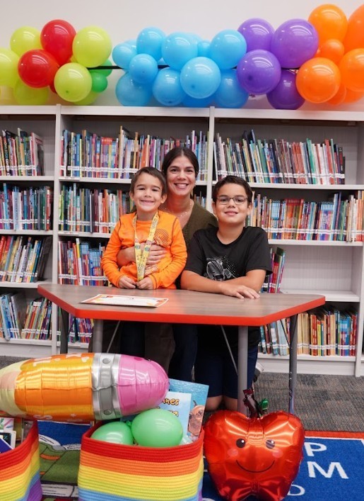 A woman and two children at a table in a library. A colorful balloon arch and books on shelves in the background.