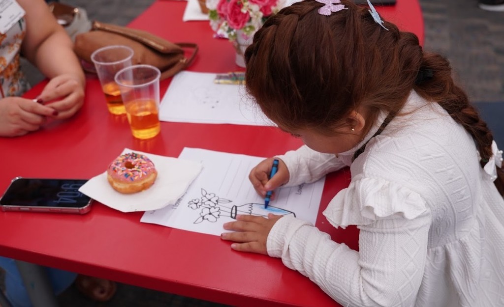 A young girl with long hair draws on a paper at a red table with two drinks and a napkin.