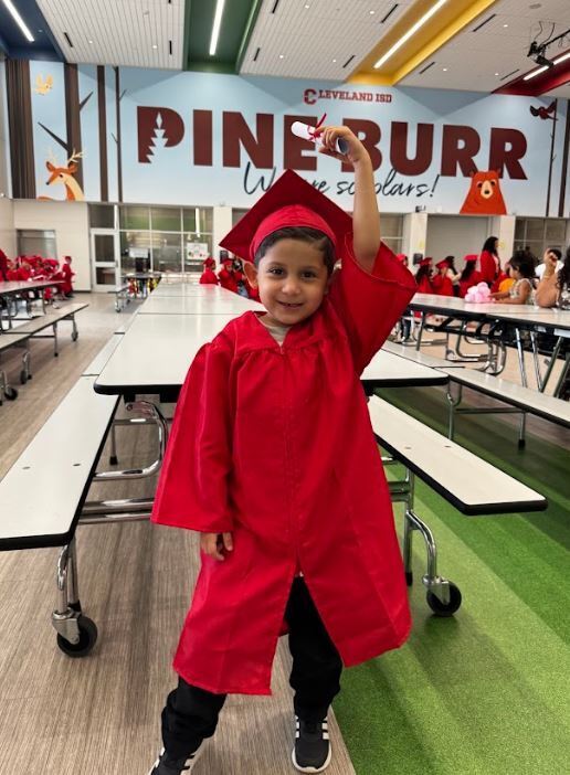 A child in a red graduation gown and cap poses for a photo in a dining hall.