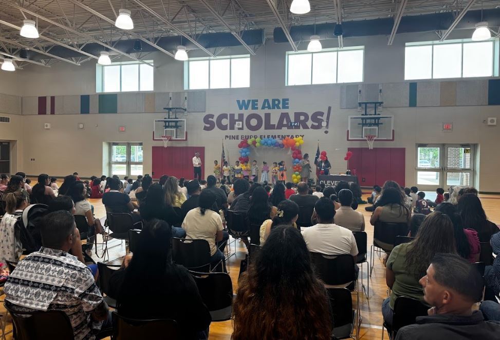 Gymnasium with a banner reading "We Are Scholars!" People sit in chairs, facing a stage with people speaking.
