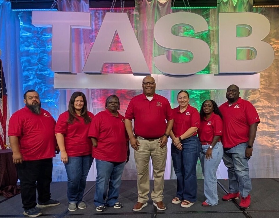 People in red shirts pose in front of a large sign with white letters that reads TASB.