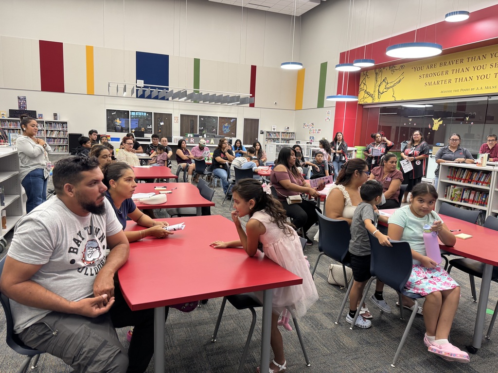 A library with red tables where a family sits, a woman stands, and a large group of children sit in chairs.