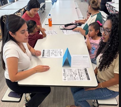 Several people seated at a table in a classroom, two women facing each other, and a child.
