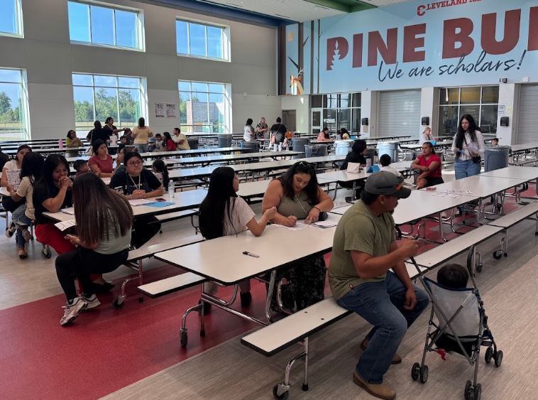 A cafeteria with multiple long tables; people sit at the tables and others walk around. Banner reads "Pine BU We are scholars!".