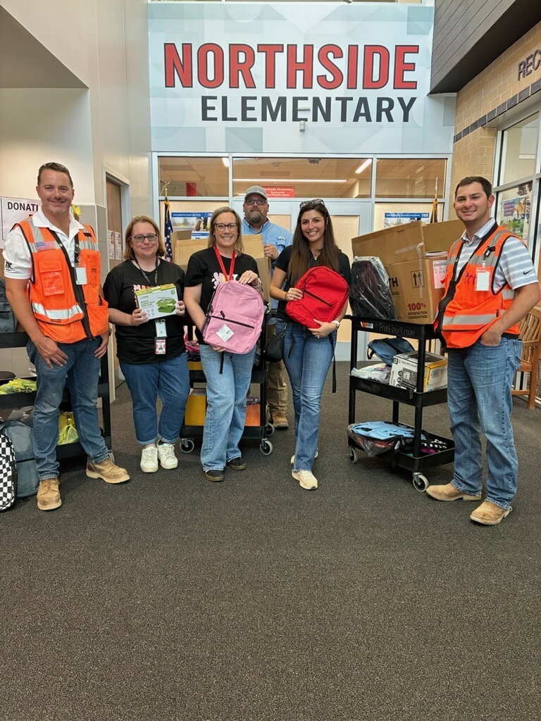 Six people in safety vests stand in front of Northside Elementary. One man holds a book, two women hold backpacks, and two others hold bags.